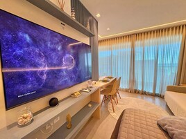 Uma sala moderna com uma televisão de tela plana grande exibindo uma cena espacial, uma área de jantar com mesa posta para dois e um espaço de estar aconchegante com uma cama.