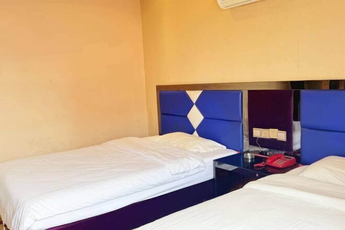 A hotel room with two single beds, a blue headboard, a bedside table with a red telephone, and a wall-mounted air conditioner.