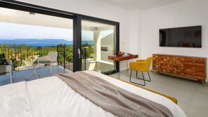 Ein modernes Schlafzimmer mit Bett, Schreibtisch und Stuhl, einem an der Wand montierten Fernseher und einem Balkon mit Blick auf Meer und Berge.