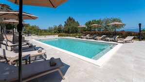 Ein rechteckiger Pool mit Liegestühlen und Sonnenschirmen, umgeben von einer Steindeckfläche und Grünflächen.