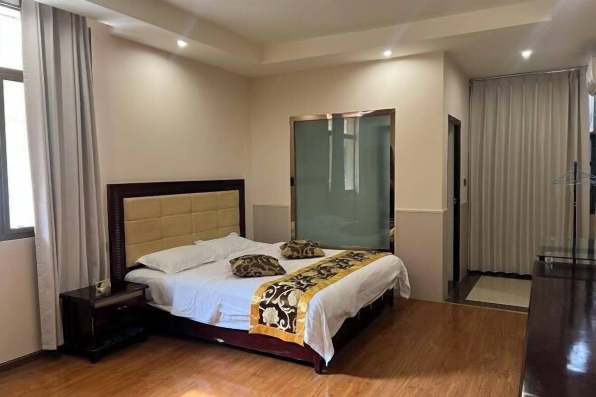 A hotel room with a large bed, a wooden nightstand, a glass partition wall, and a view of the outside.