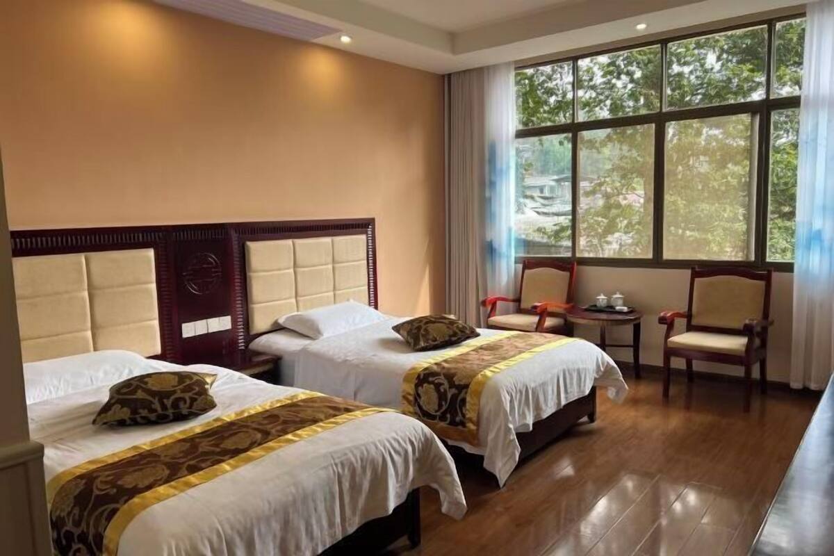 A hotel room with two beds, a small table, a chair, and a large window with a view of trees.
