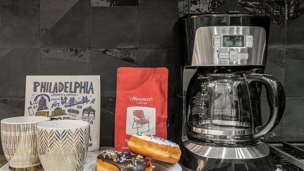 Una encimera de cocina con una cafetera, dos tazas decorativas, un paquete de café y un plato con una rosquilla.