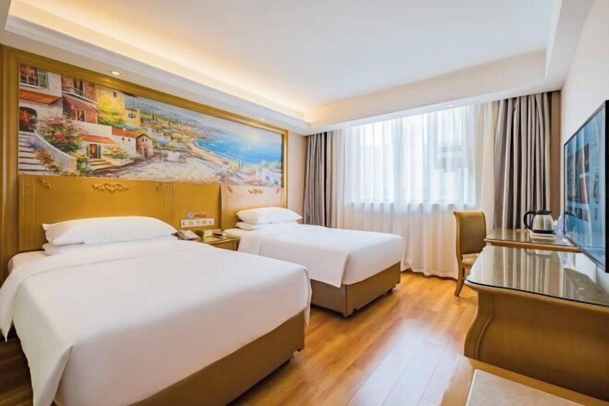 A hotel room with two beds, a painting on the wall, a dining table, and a view of the outdoors.