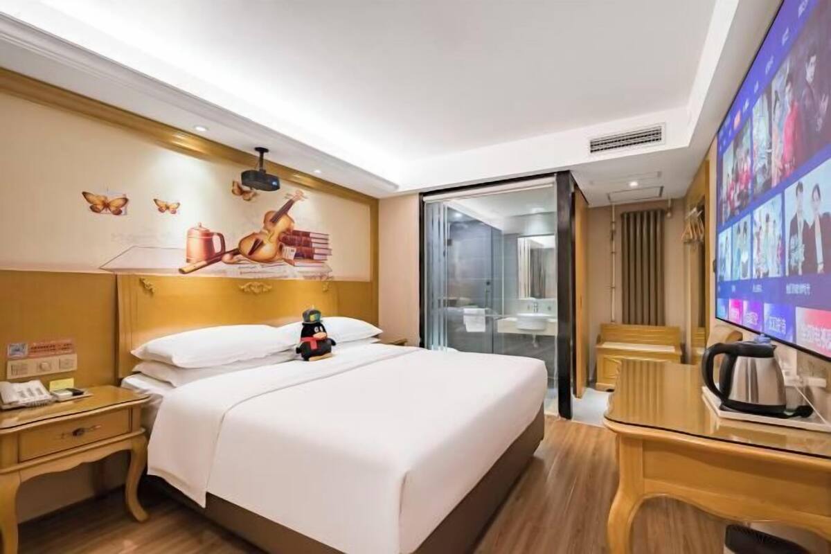 A hotel room with a bed, bedside table, a television, and a glass partition leading to a bathroom.