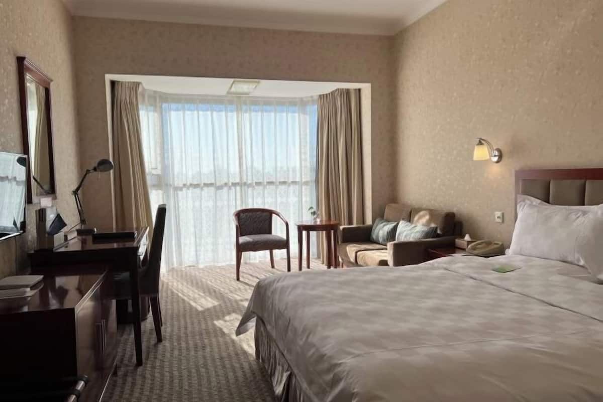 A hotel room with a large bed, a desk with a chair, a small table, and a sofa. There is a window with curtains and a lamp on the wall.
