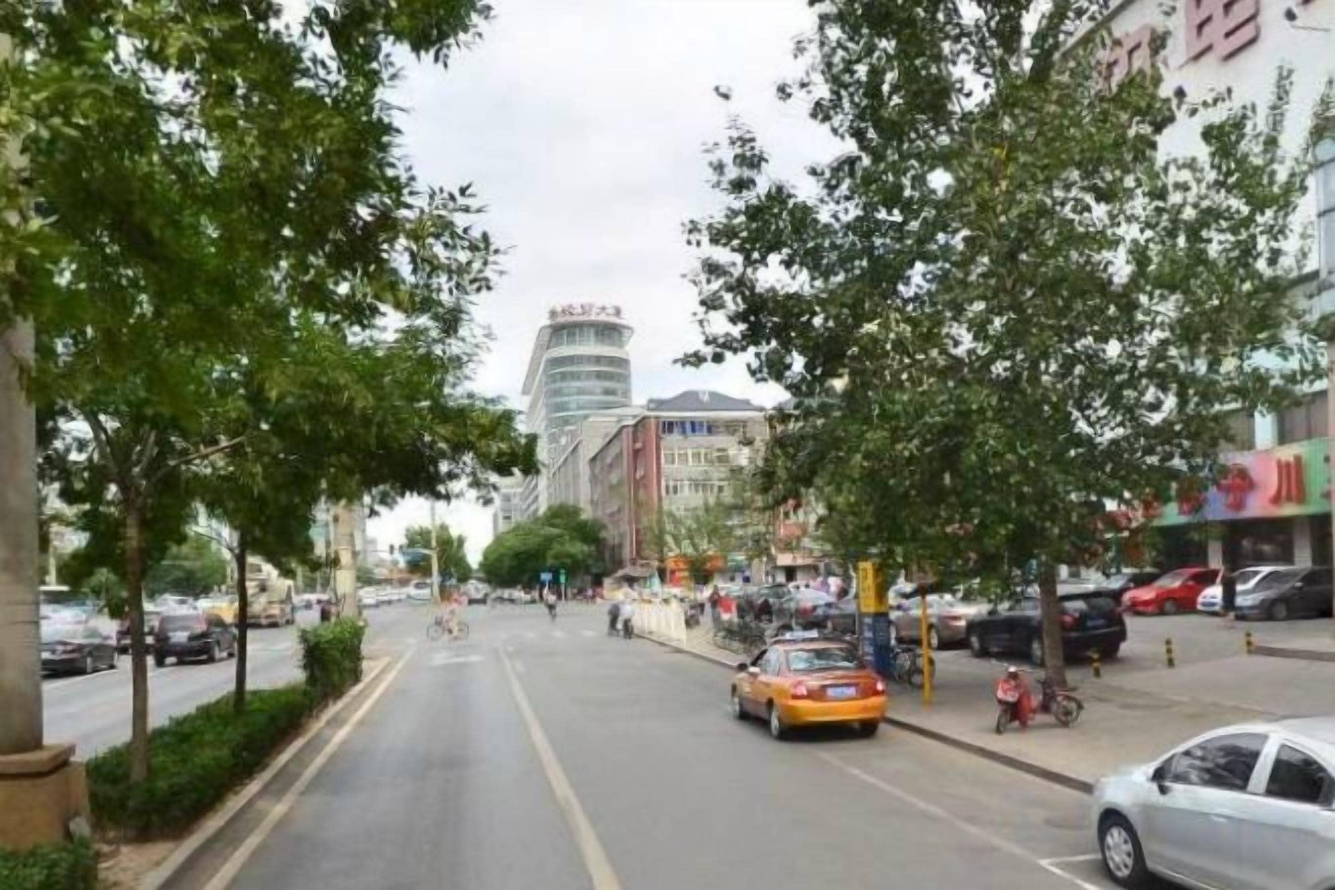 A street with cars, a yellow taxi, and pedestrians. There are trees lining the road, and buildings in the background.
