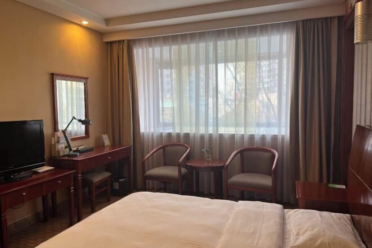 Quarto de hotel com cama, escrivaninha com computador, televisão, duas cadeiras e uma janela grande com cortinas.