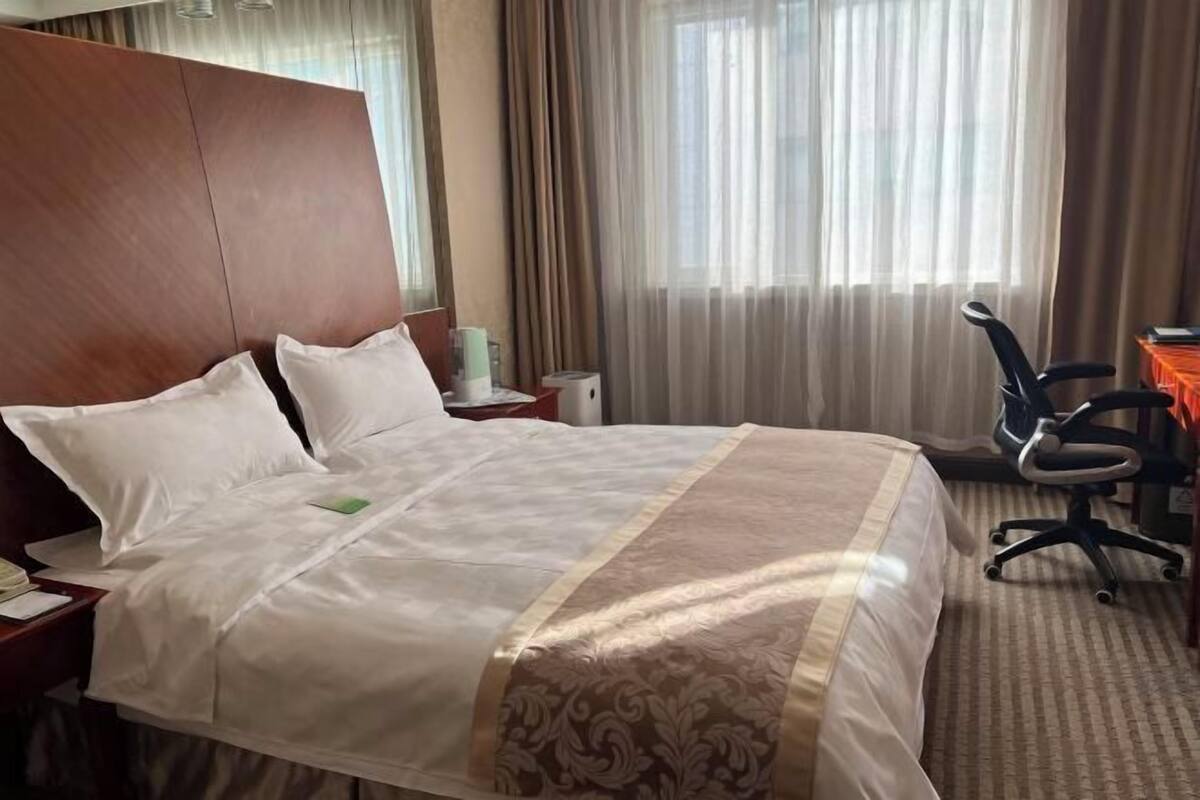 A hotel room with a large bed, a desk with a chair, a nightstand, and a window with curtains.