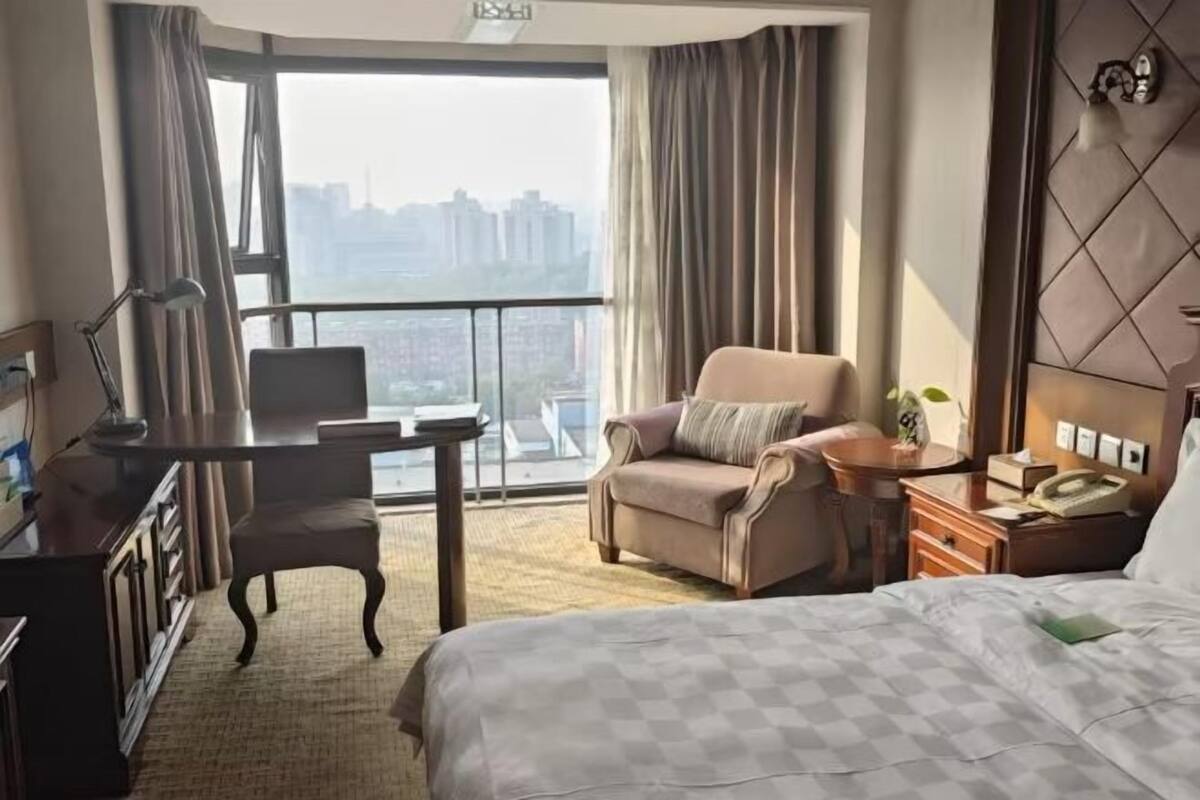 A hotel room with a bed, a desk, a chair, a nightstand, and a view of a cityscape through large windows.
