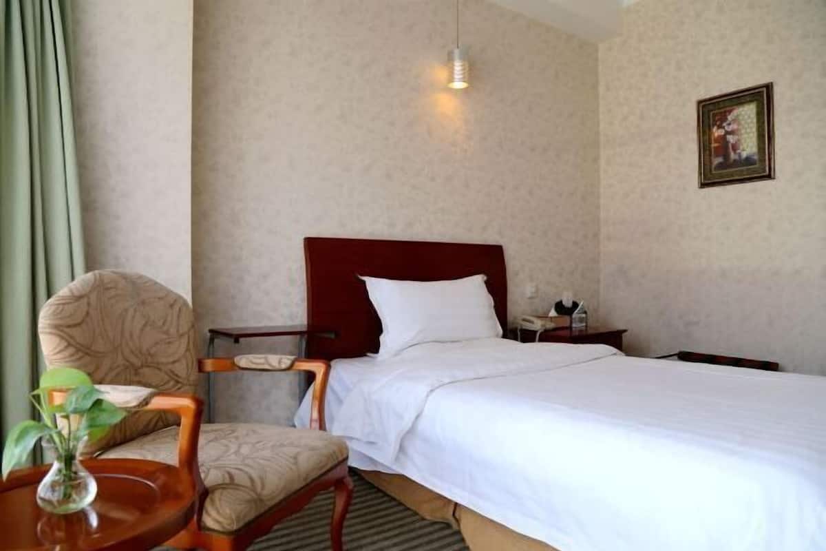 A hotel room with a bed, a chair, a small table with a vase, a nightstand, and a hanging light.