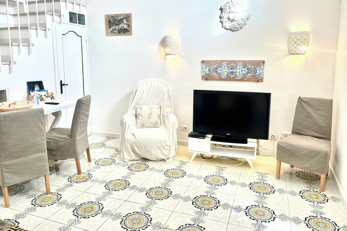 A living room with a TV, two chairs, a white sofa, patterned floor tiles, a staircase, and wall-mounted lights.