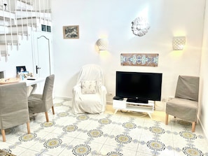 A living room with a TV, two chairs, a white sofa, patterned floor tiles, a staircase, and wall-mounted lights.