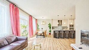 Una sala de estar moderna con un sofá de cuero, piso de madera, un comedor con mesa y sillas, una cocina con taburetes de bar y un televisor sobre un mueble.