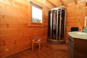 Ein Badezimmer einer Holzhütte mit einer Glasdusche, einem hölzernen Hocker und einem Waschbecken mit Spiegel.