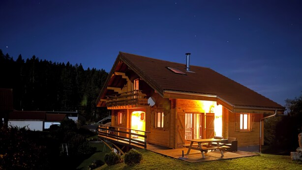 A wooden cabin with a balcony, a chimney, and a well-lit interior. - Chalet for 9 people. Skiing, Hiking, and Nature - Haut-Jura (Prémanon)