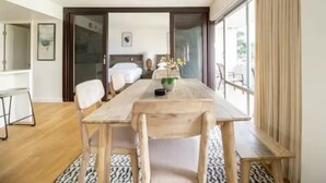 Une chambre d’hôtel moderne avec une table à manger en bois, des chaises blanches, une plante en pot et une vue sur l’extérieur à travers une porte vitrée.