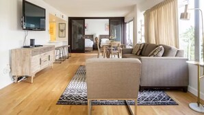 Un salon moderne avec un canapé, un téléviseur fixé au mur, un coin repas comprenant une table et des chaises, et une vue sur la chambre à coucher à travers une porte ouverte.