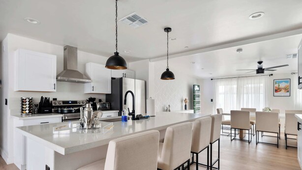 A modern kitchen with white cabinetry, a central island with bar stools, and a dining area with a high ceiling fan.