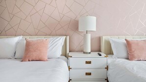 A hotel room with two beds, each with white bedding and pink pillows, a nightstand with a lamp, and a wall with a geometric pattern.