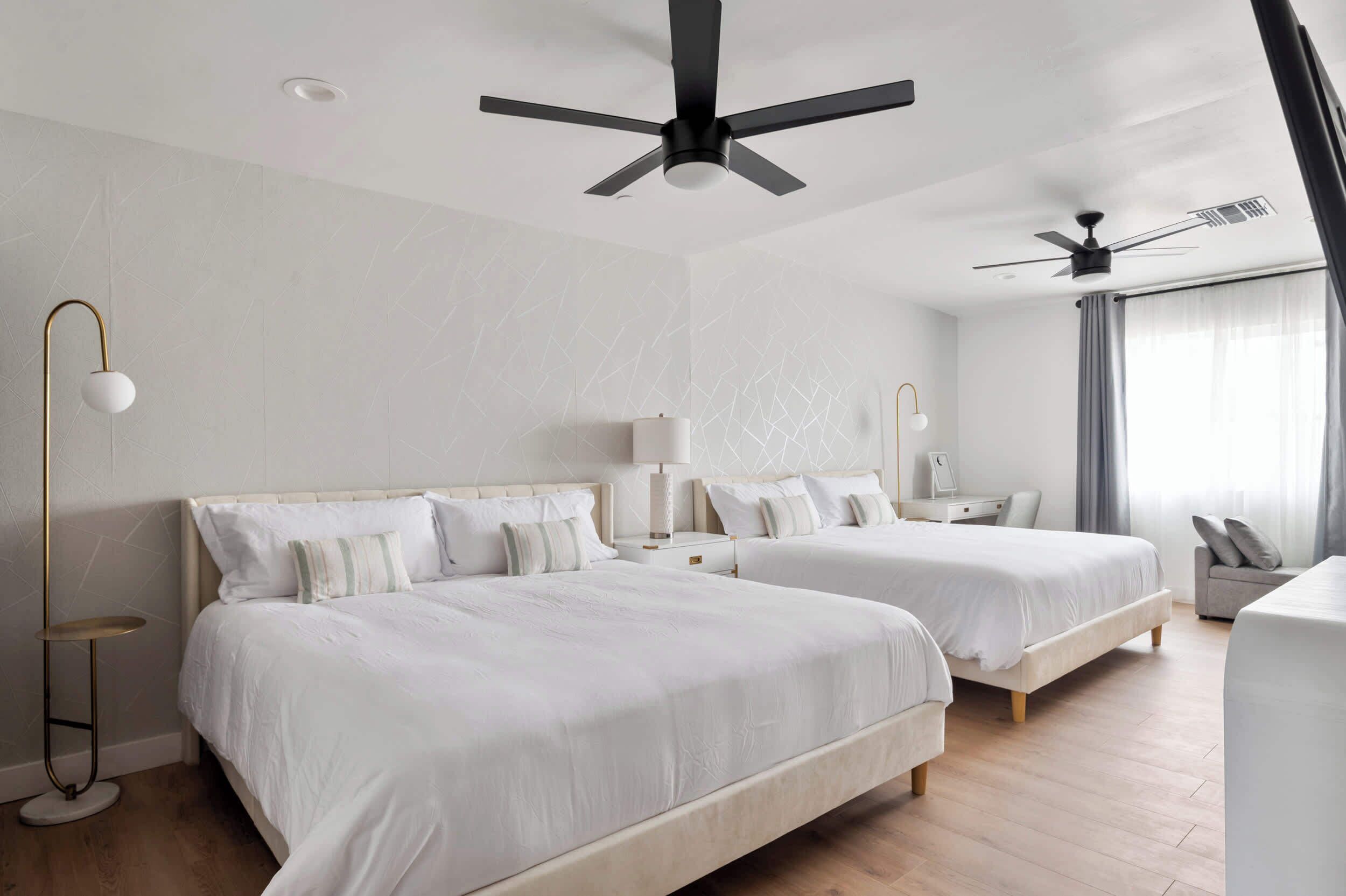 Habitación de hotel con dos camas, ventilador de techo, escritorio, silla y ventana con cortinas.