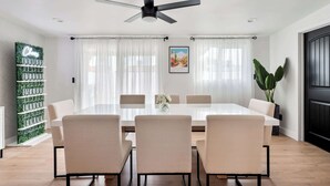 A modern dining room with a white table, beige chairs, a ceiling fan, and a large window with sheer curtains.