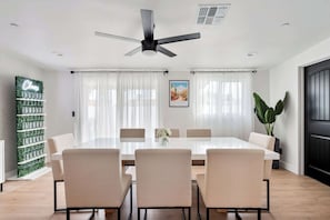 A modern dining room with a white table, beige chairs, a ceiling fan, and a large window with sheer curtains.