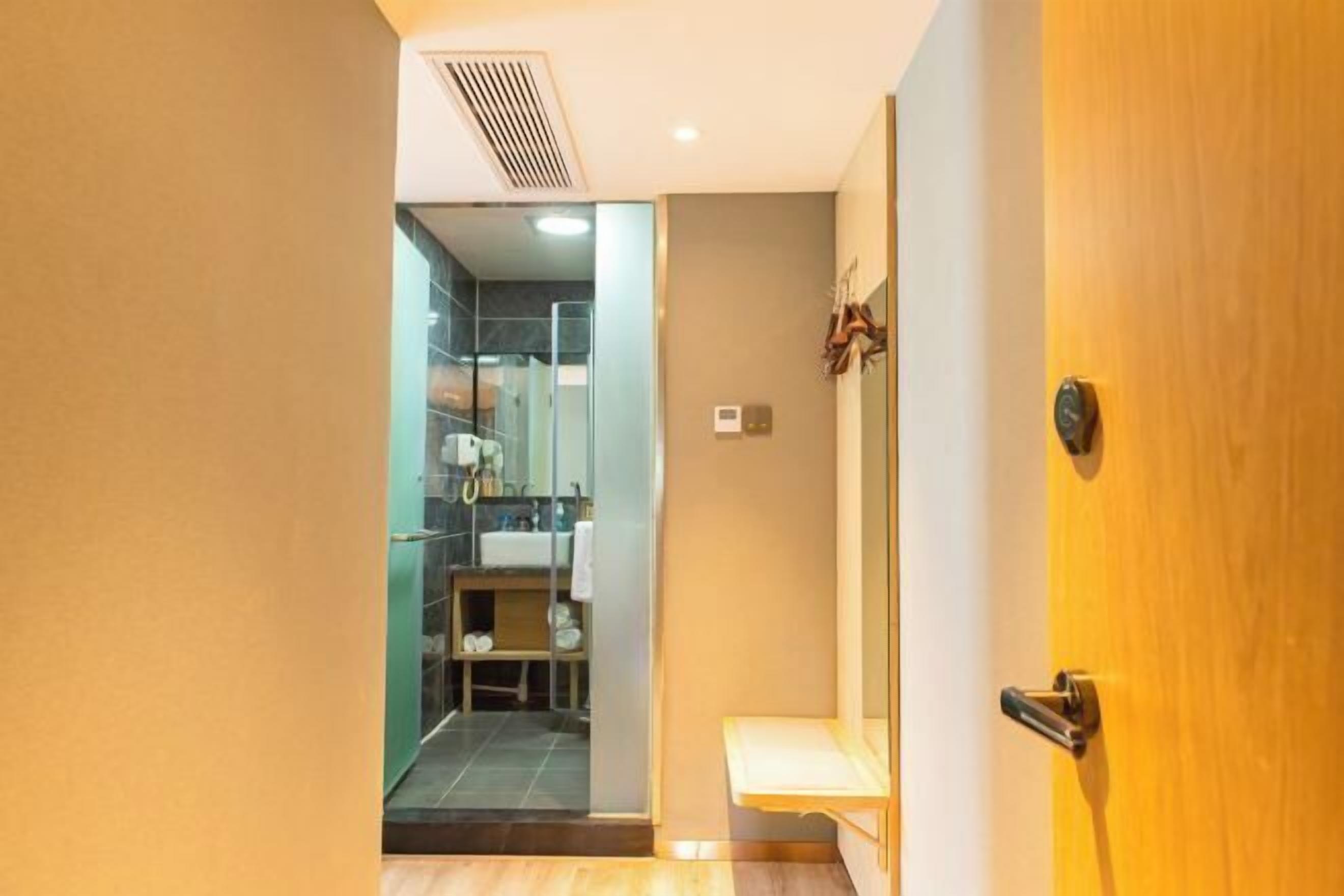 Quarto de hotel com porta de madeira, banheiro pequeno com chuveiro de vidro e um banco de madeira.