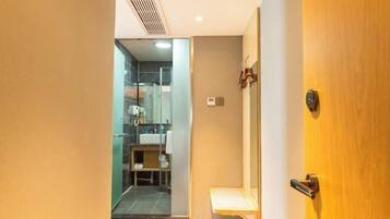 Quarto de hotel com porta de madeira, banheiro pequeno com chuveiro de vidro e um banco de madeira.