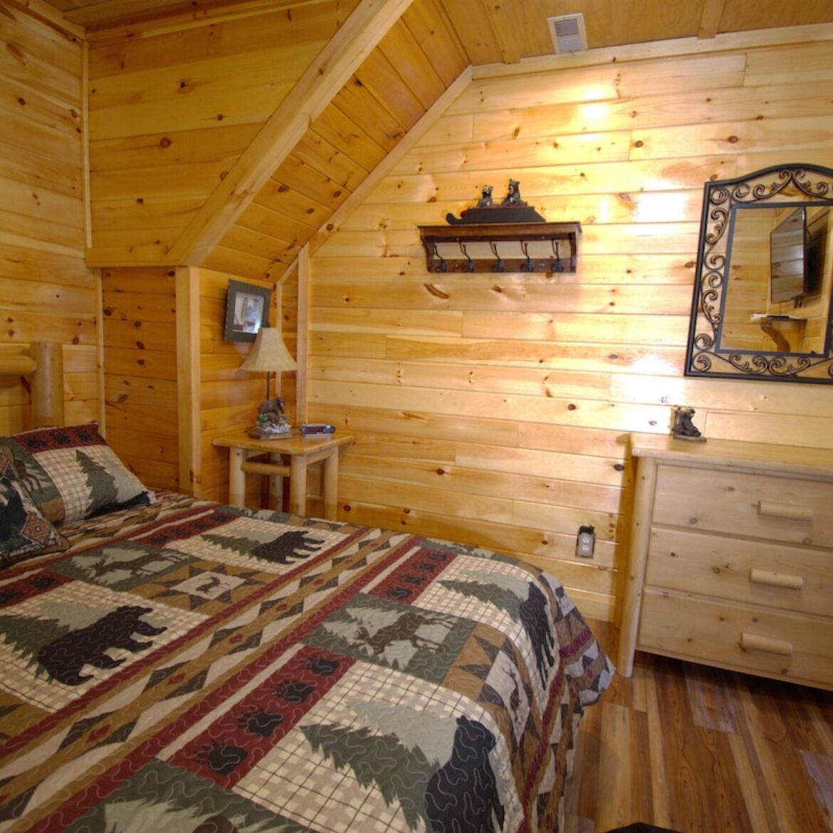 A log cabin bedroom with a bed, dresser, mirror, lamp, and a shelf with decorative items.