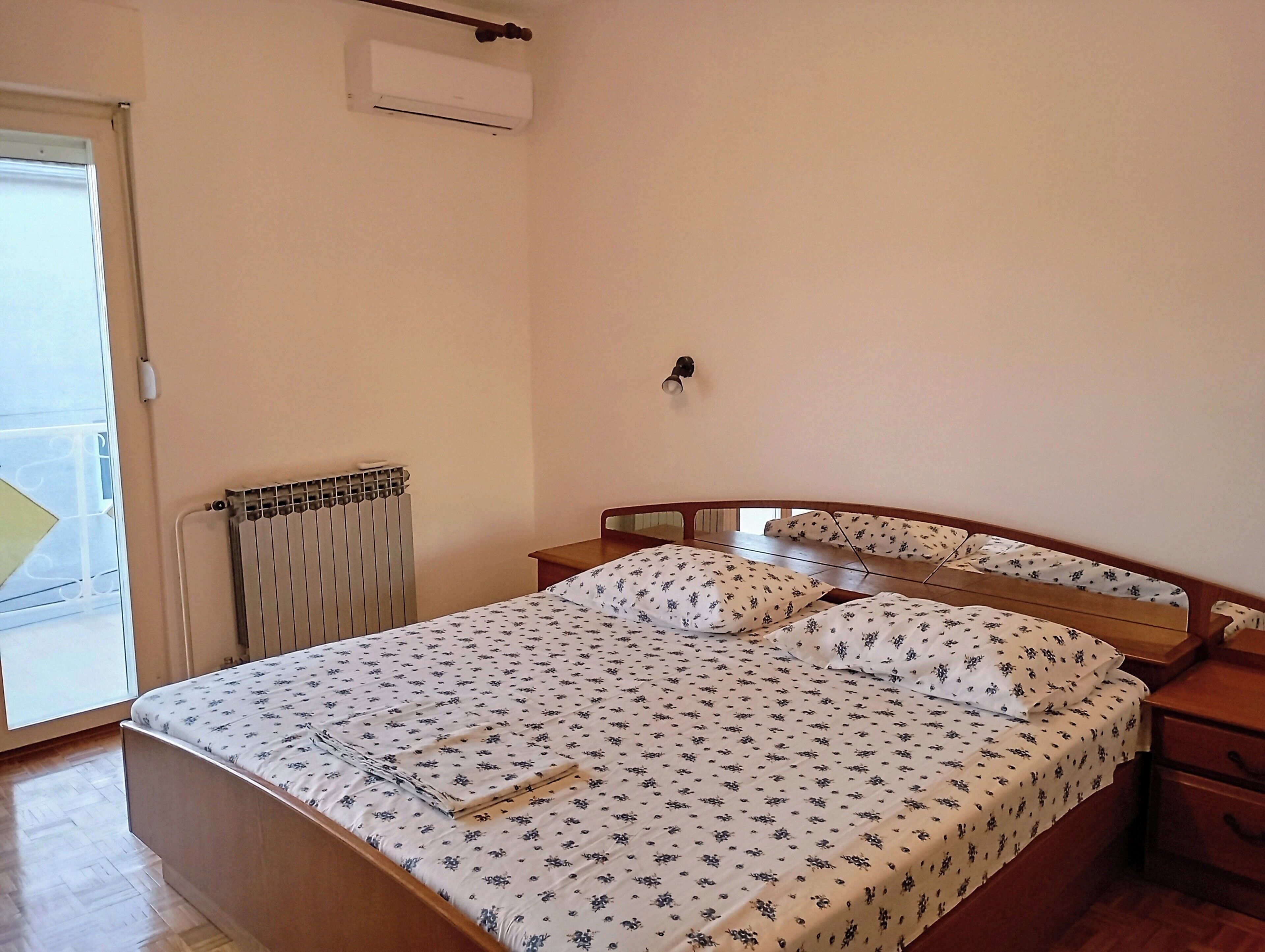 A bedroom with a bed, a radiator, a wall-mounted air conditioner, and a window with a view.