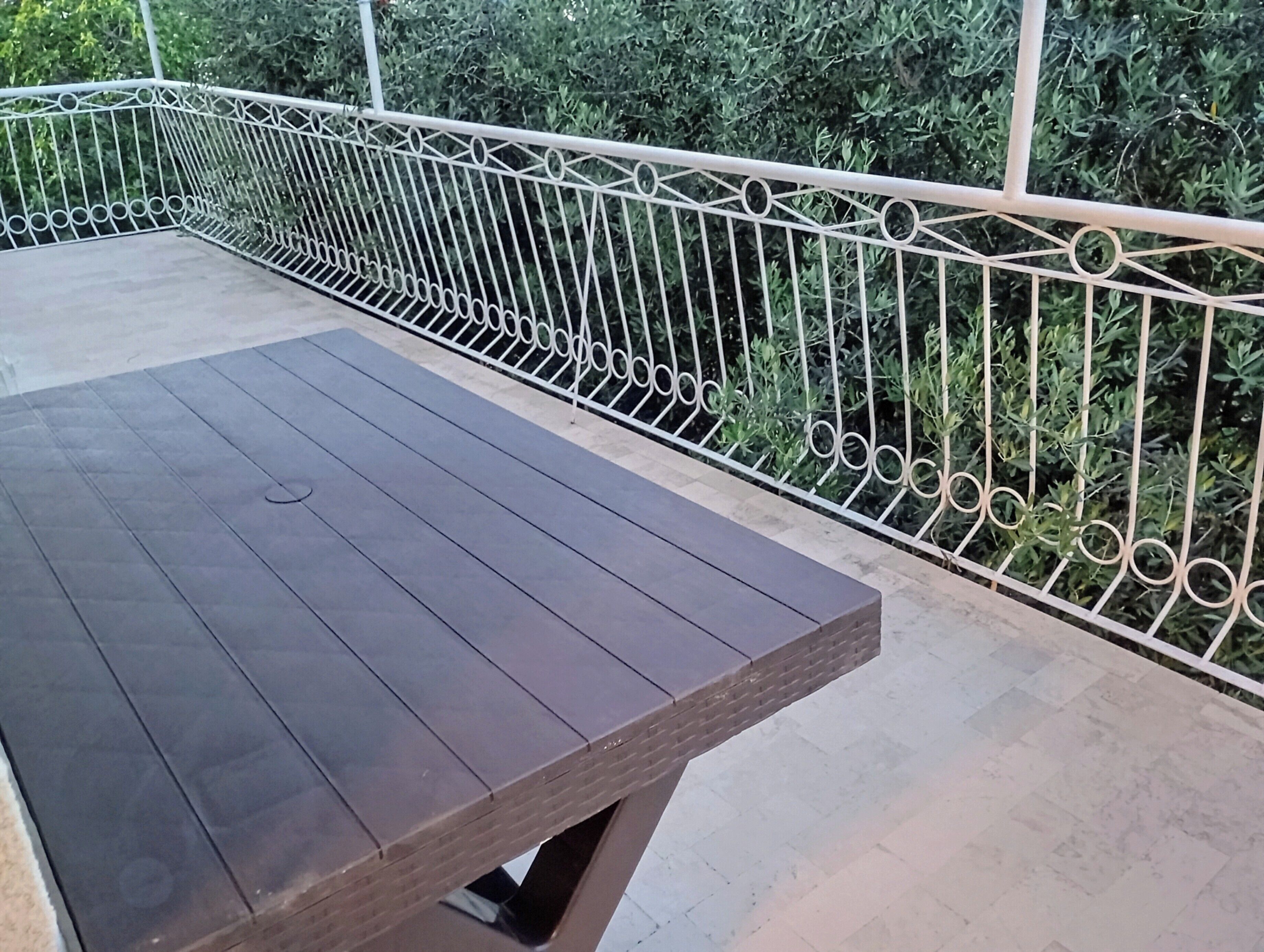 A wooden bench on a balcony with a white metal railing and greenery in the background.