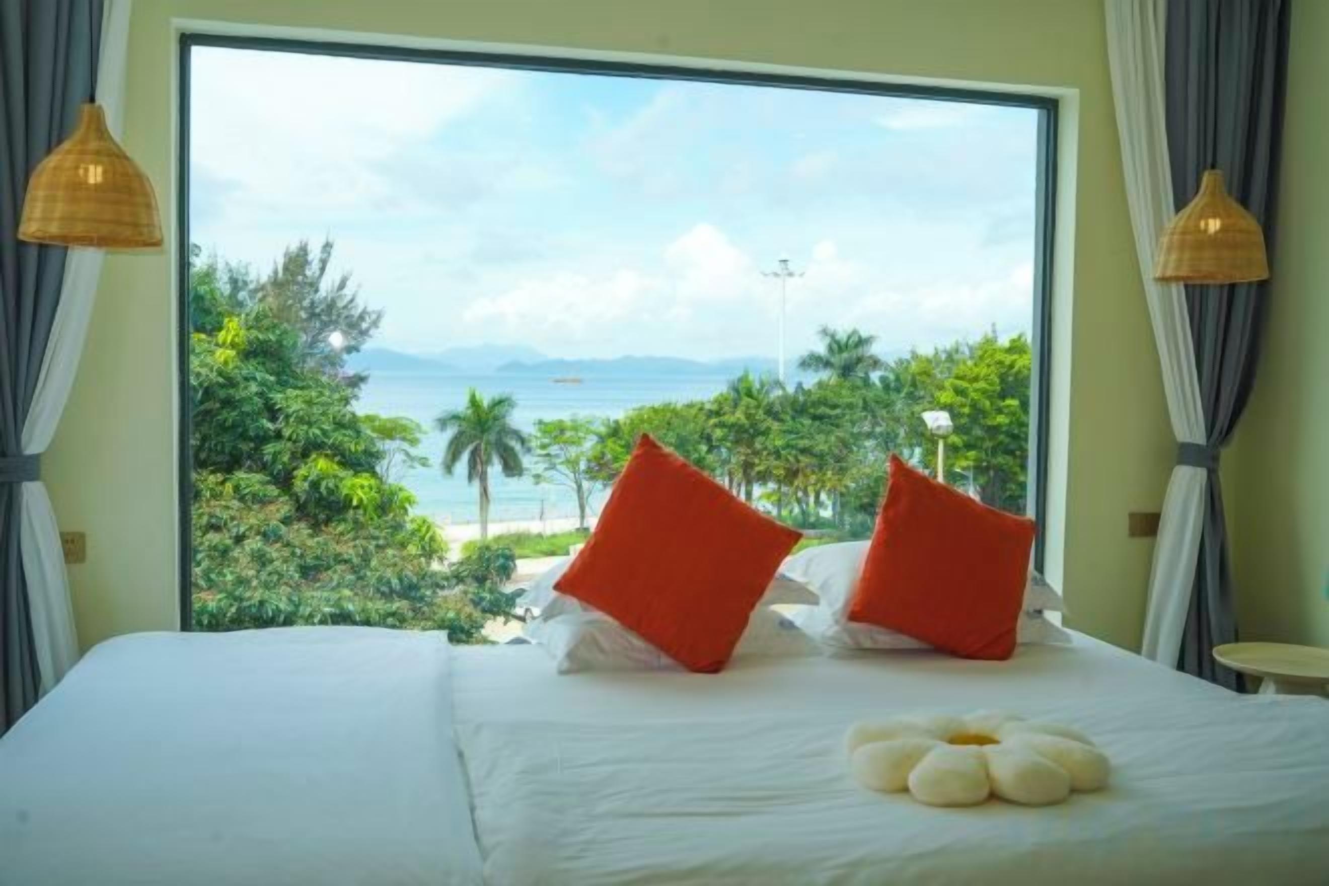 A bed with a view of a beach and palm trees, a window with curtains, and a small table with a lamp.