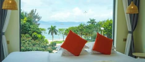 A bed with a view of a beach and palm trees, a window with curtains, and a small table with a lamp.