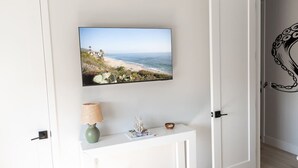 A room with a flat-screen TV mounted on the wall displaying a beach scene, a small console table with a lamp, a vase, and a bowl, and a door with a decorative octopus mural.