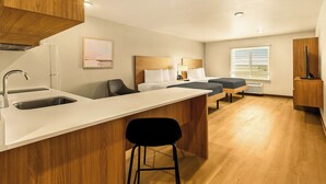 A modern kitchen with a wooden countertop, a sink, and a bar stool. There are two beds with white linens and a window with blinds. - LivAway Suites Seattle-Renton (Renton)