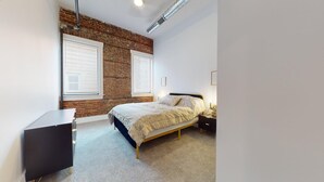 A bedroom with a brick wall, two windows with blinds, a bed with a headboard, a bedside table, and a desk.