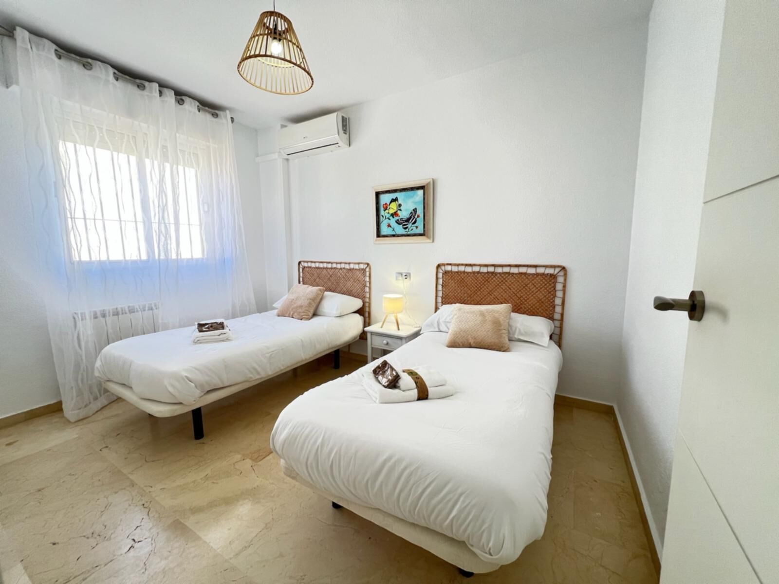 A hotel room with two single beds, a small bedside table with a lamp, a window with sheer curtains, and a wall-mounted air conditioner.