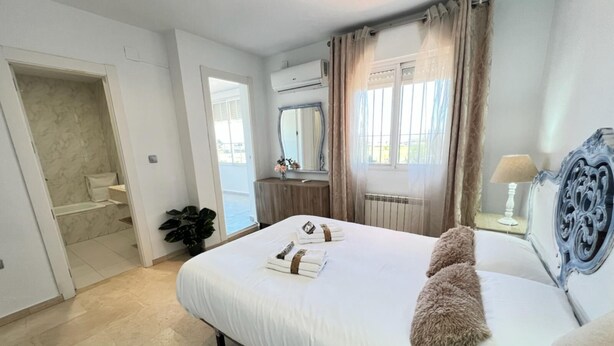 Un dormitorio con una cama grande, una mesita de noche con lámpara, un espejo y vistas al exterior a través de una ventana con cortinas.