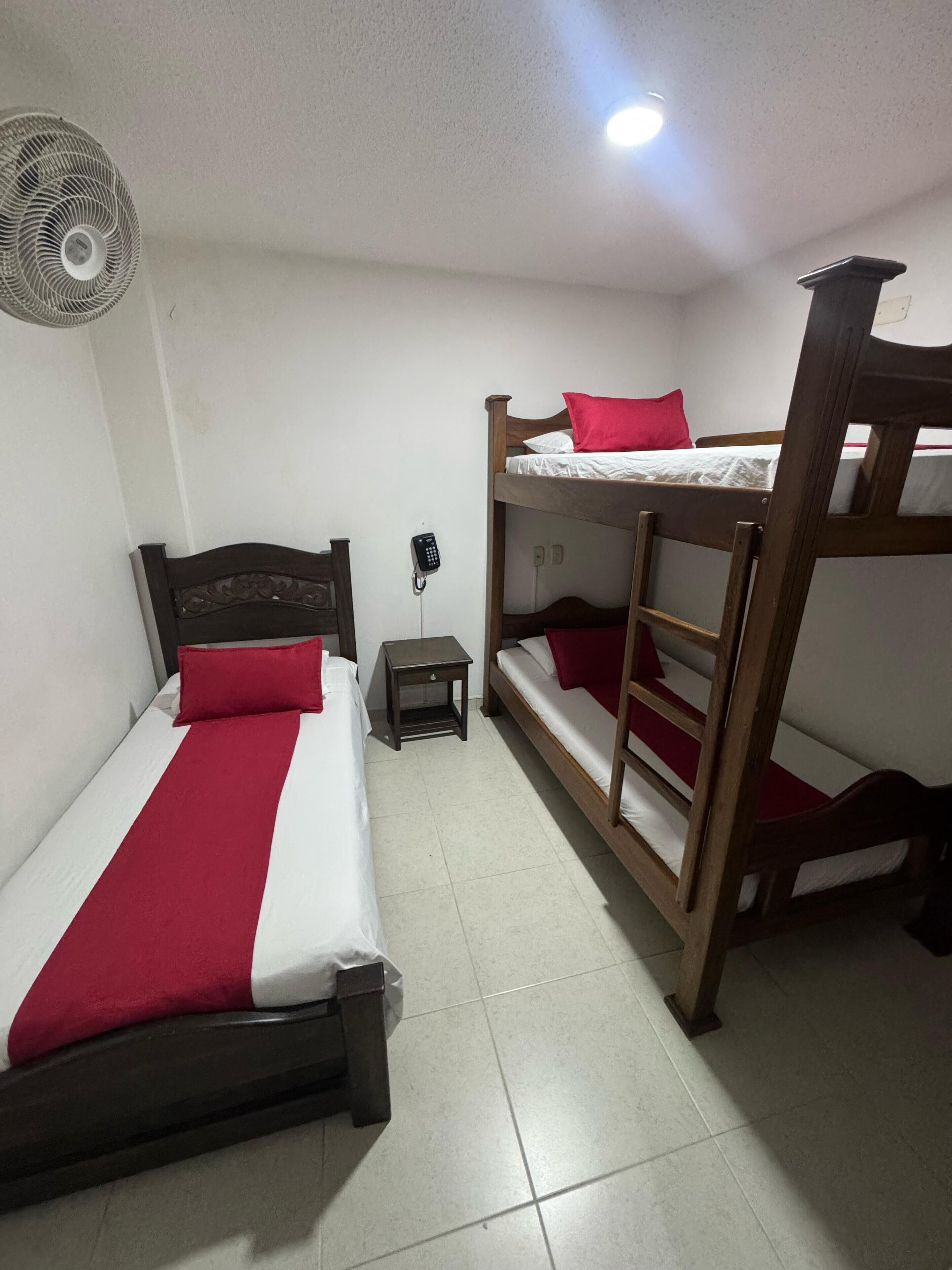 A room with two beds, one with a red and white bedspread and another with a wooden bunk bed. There is a small table with a phone and a fan on the wall.
