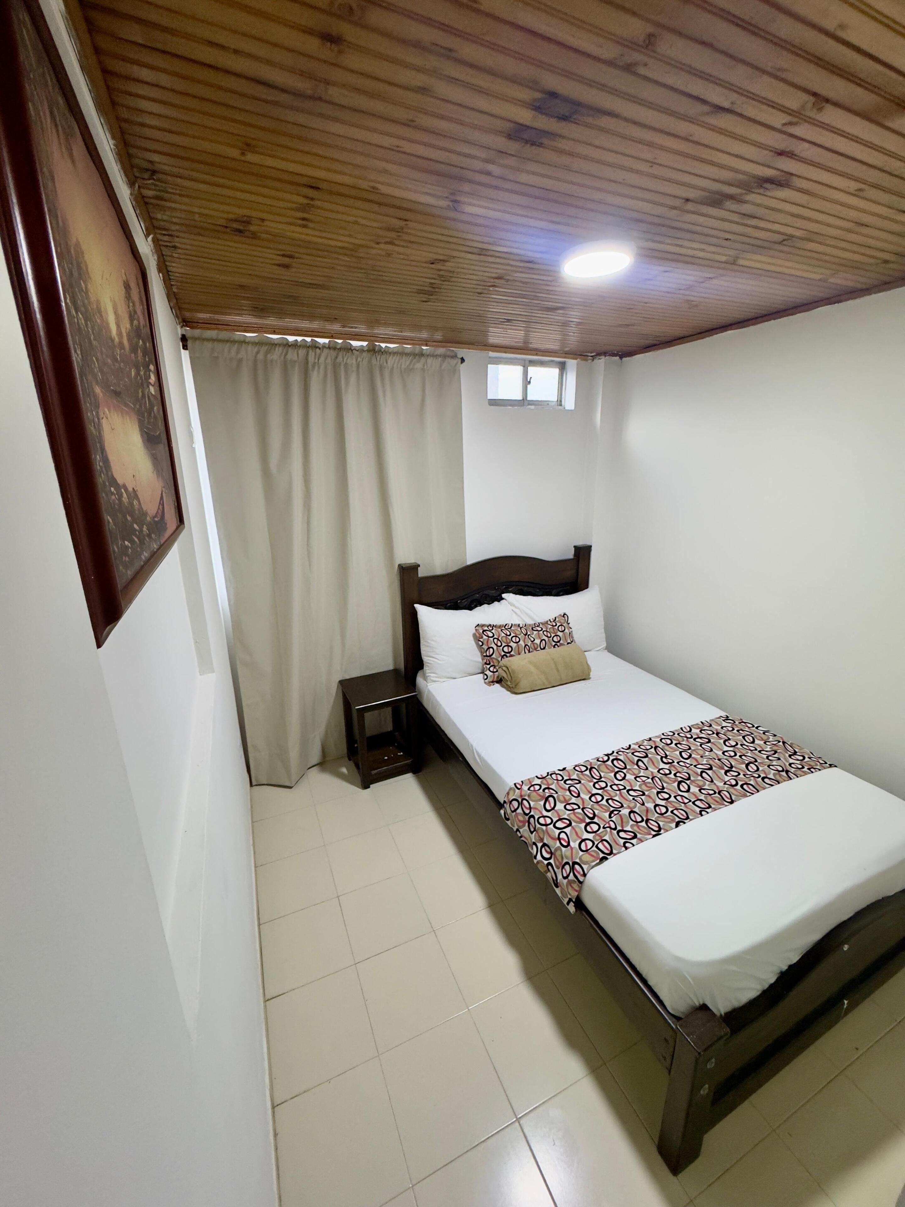 A bedroom with a wooden ceiling, a bed with a patterned bedspread, a nightstand, a window with curtains, and a painting on the wall.