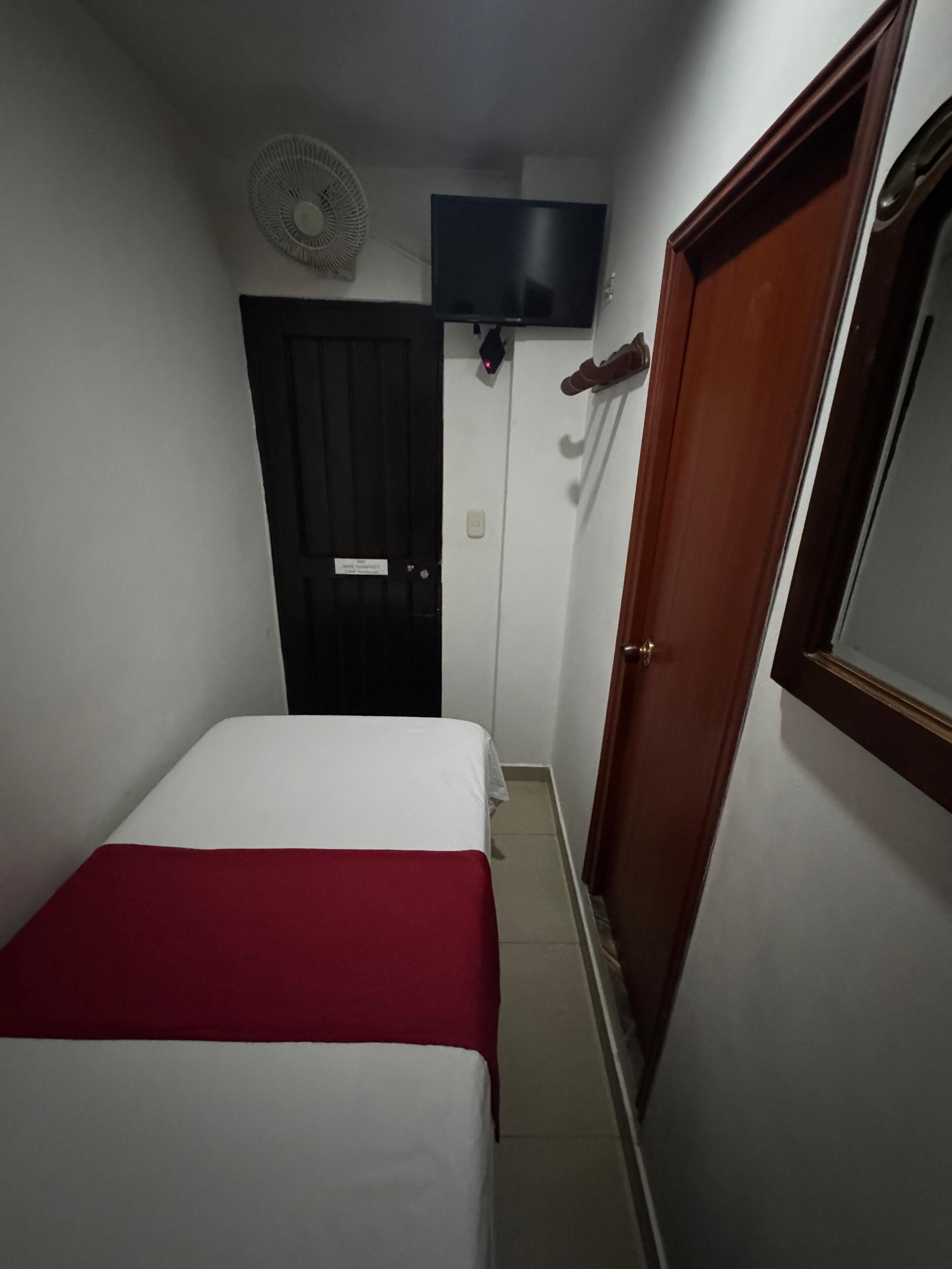 Una habitación de hotel pequeña y estrecha con una cama, una puerta de madera, un ventilador de pared y un televisor.