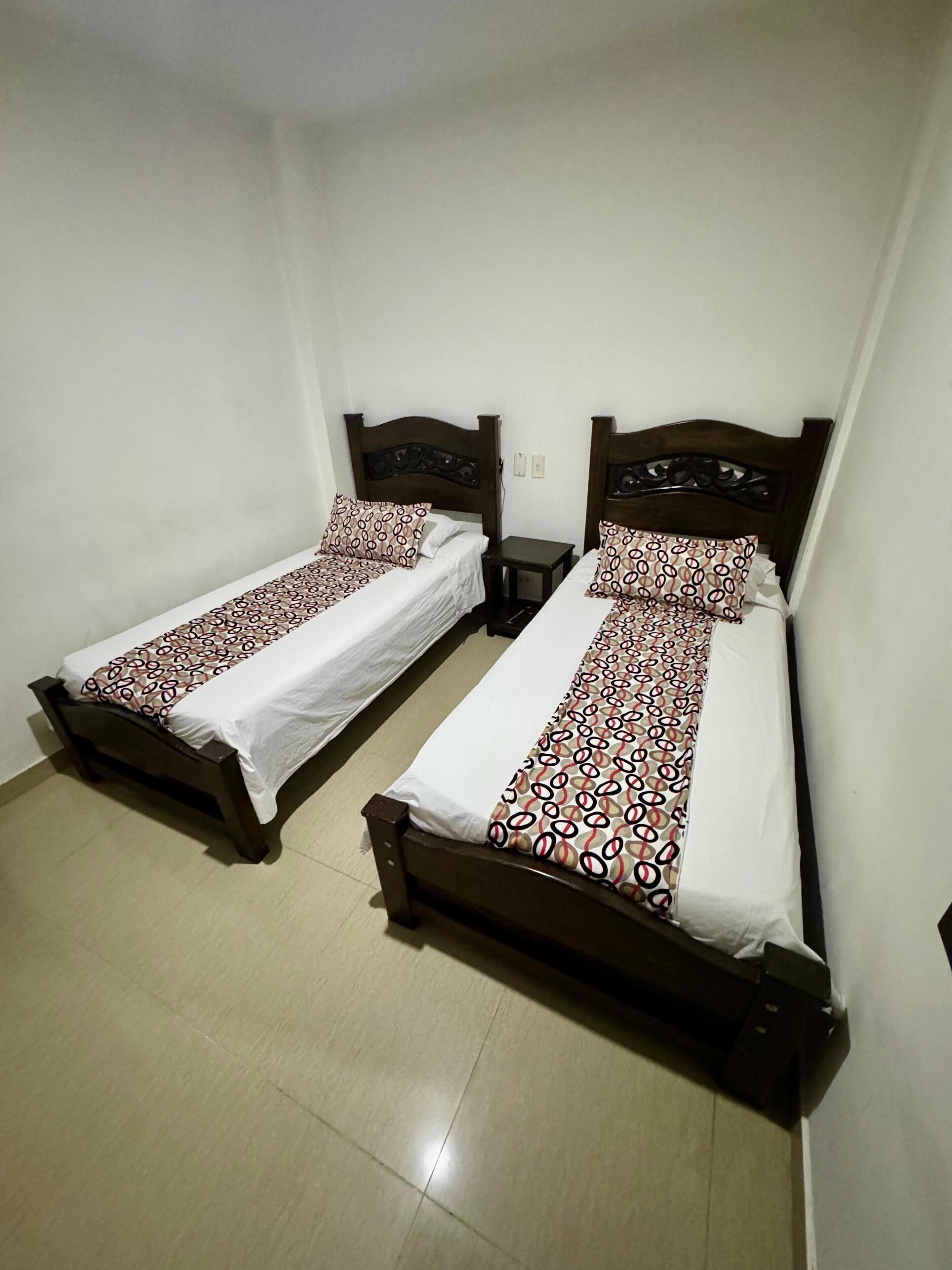 Dos camas individuales con cabeceros de madera y colchas estampadas en una habitación con paredes blancas y suelo de baldosas.