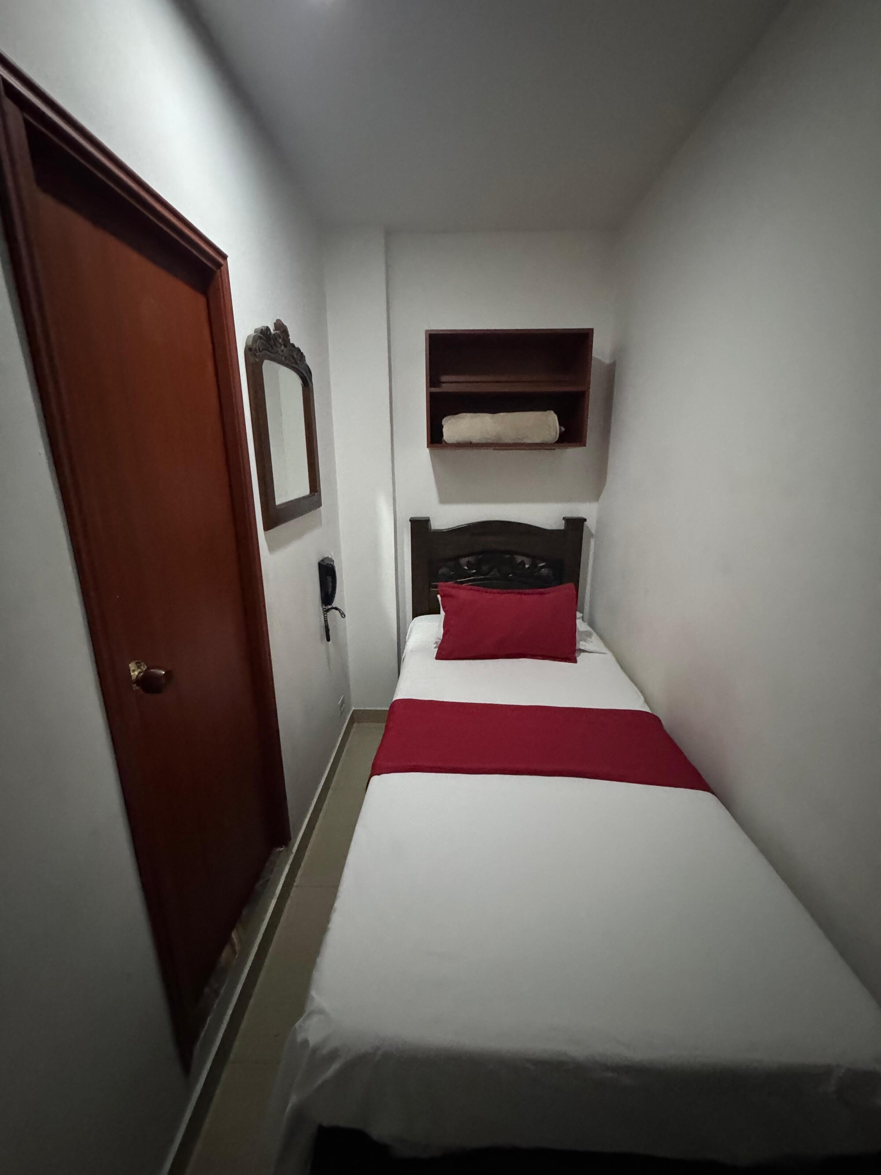 A small, narrow hotel room with a single bed, a wooden door, a bedside table, and a shelf above the bed.