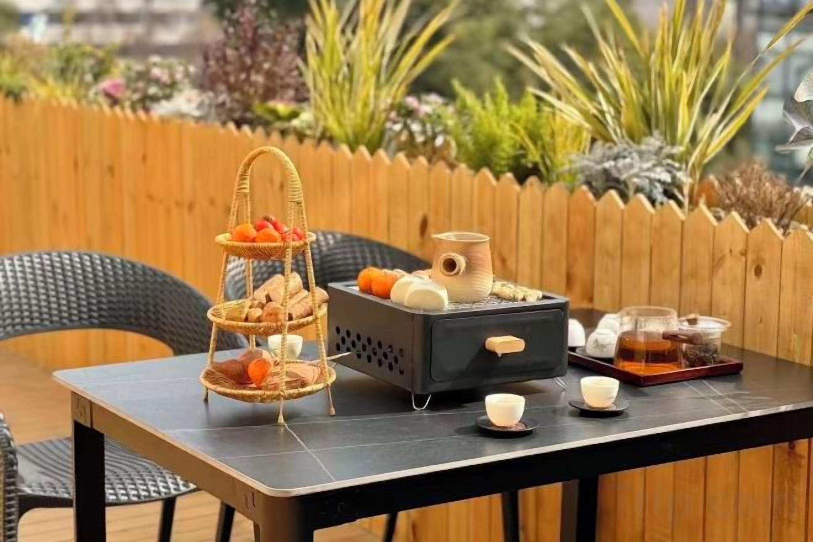 Mesa al aire libre con una bandeja de tres niveles para fruta y pan, una cafetera y una bandeja con tazas y tetera.