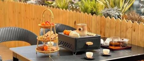 A table set outdoors with a three-tiered fruit and bread display, a coffee pot, and a tray with cups and a teapot.