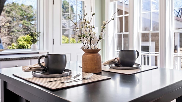 Una mesa de comedor con dos tazas y platillos negros, un florero de madera con flores, y una ventana con marcos blancos.