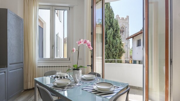 A dining area with a glass table set for two, a potted orchid, and a view of a building with a tower through a sliding glass door. - Modern Comfort in Florence (Firenze)