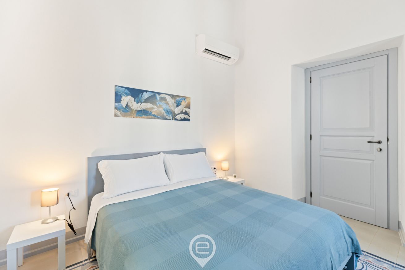 A bedroom with a bed, bedside lamps, an air conditioner, a door, and a painting on the wall.