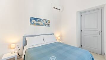 A bedroom with a bed, bedside lamps, an air conditioner, a door, and a painting on the wall.